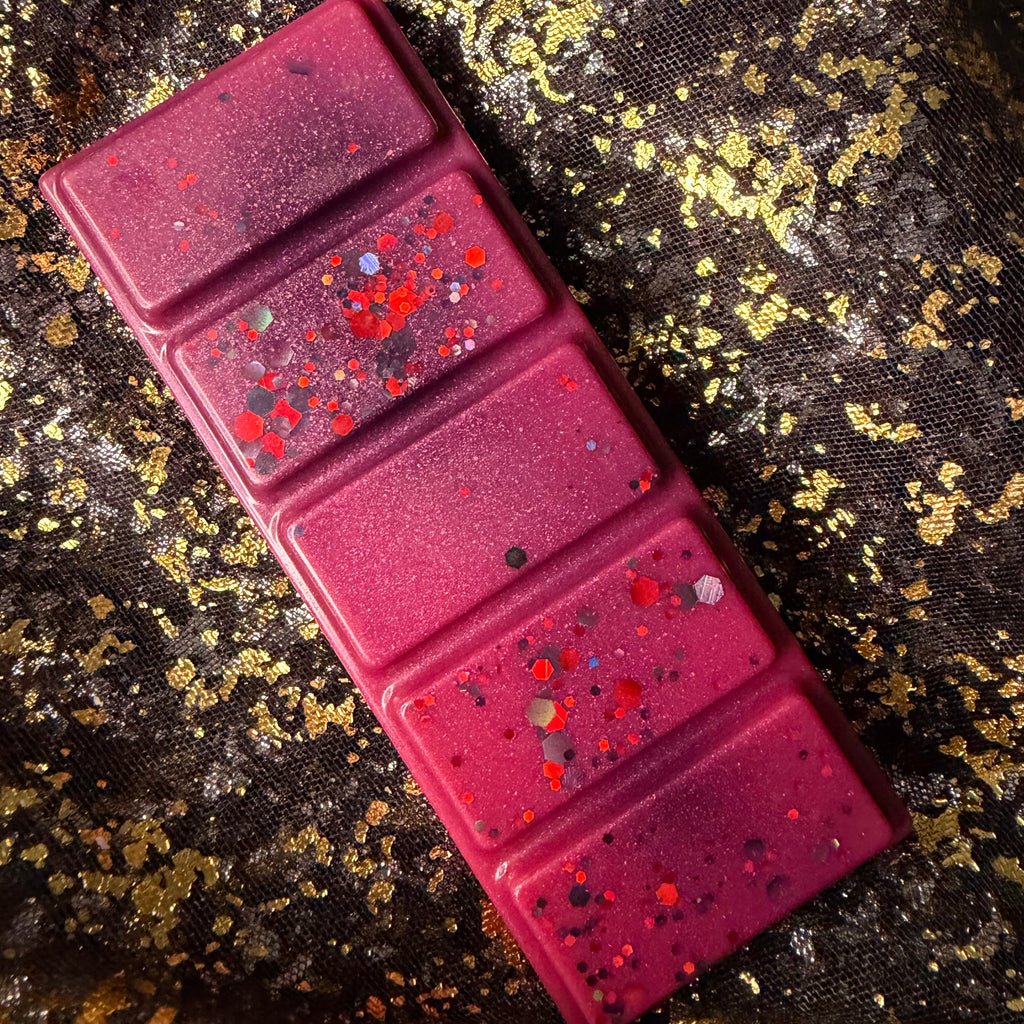 Deep magenta Theo de Fangle soy wax melt displayed diagonally on black-and-gold fabric, showing five rectangular segments decorated with red, orange and charcoal glitter.