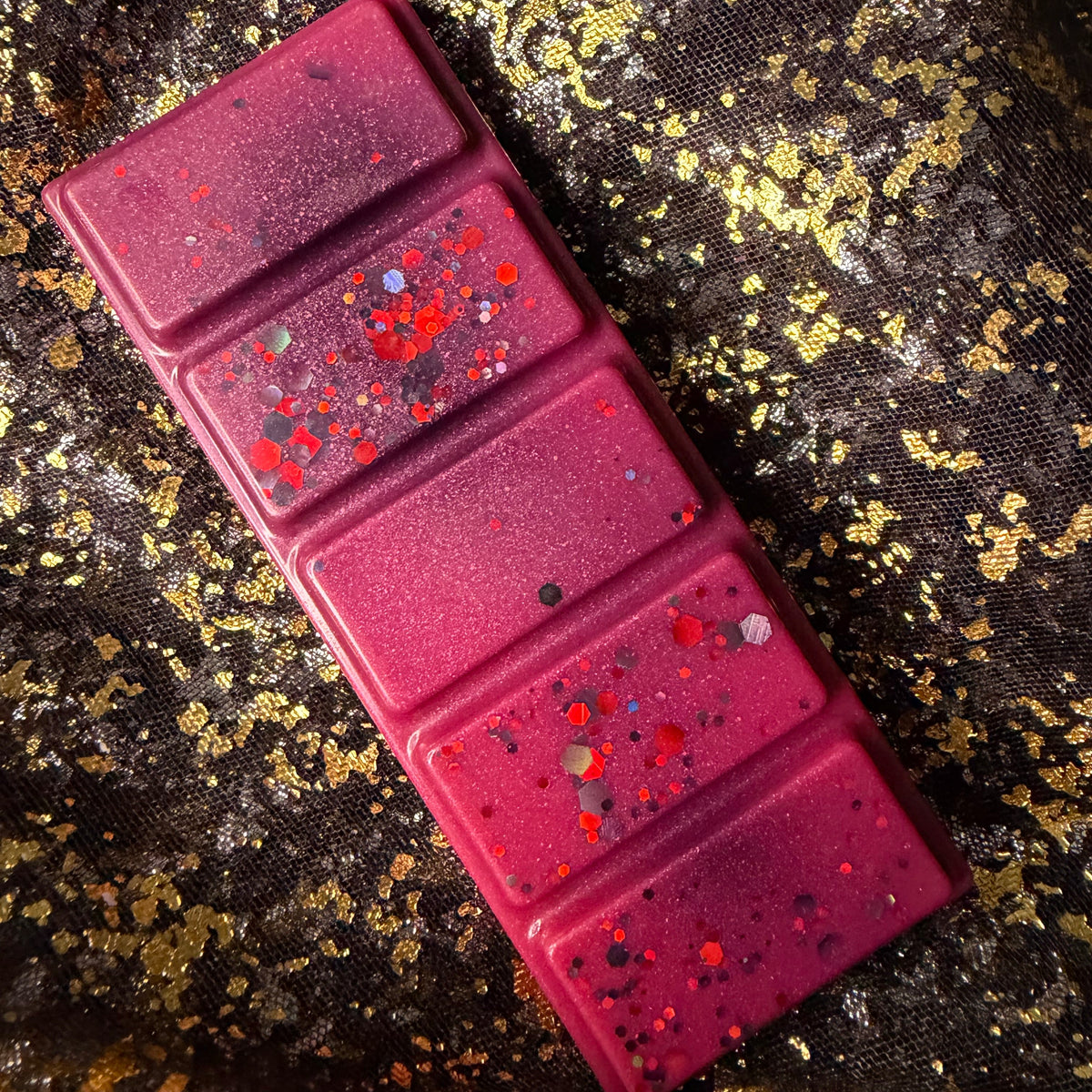 Deep magenta Theo de Fangle soy wax melt displayed diagonally on black-and-gold fabric, showing five rectangular segments decorated with red, orange and charcoal glitter.
