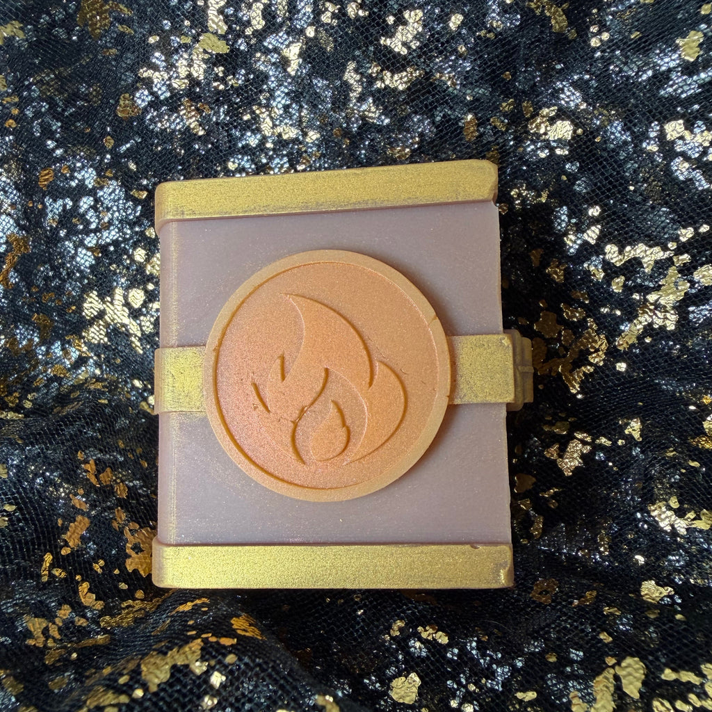 Close view of the Fire mango and vanilla scented spellbook wax melt, with an orange flame emblem between gold bands on its dusty rose cover.