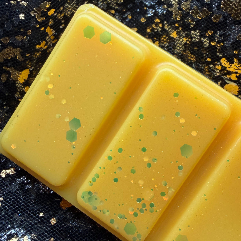 Close-up of two Summer Slow Burn wax melt segments showing fine green and gold glitter scattered across golden-yellow wax.