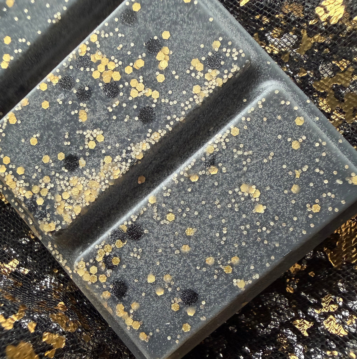 Close-up of two charcoal-black Blonde Bombshell wax melt segments decorated with fine gold glitter, larger gold hexagons and small black circular pieces against black-and-gold fabric.
