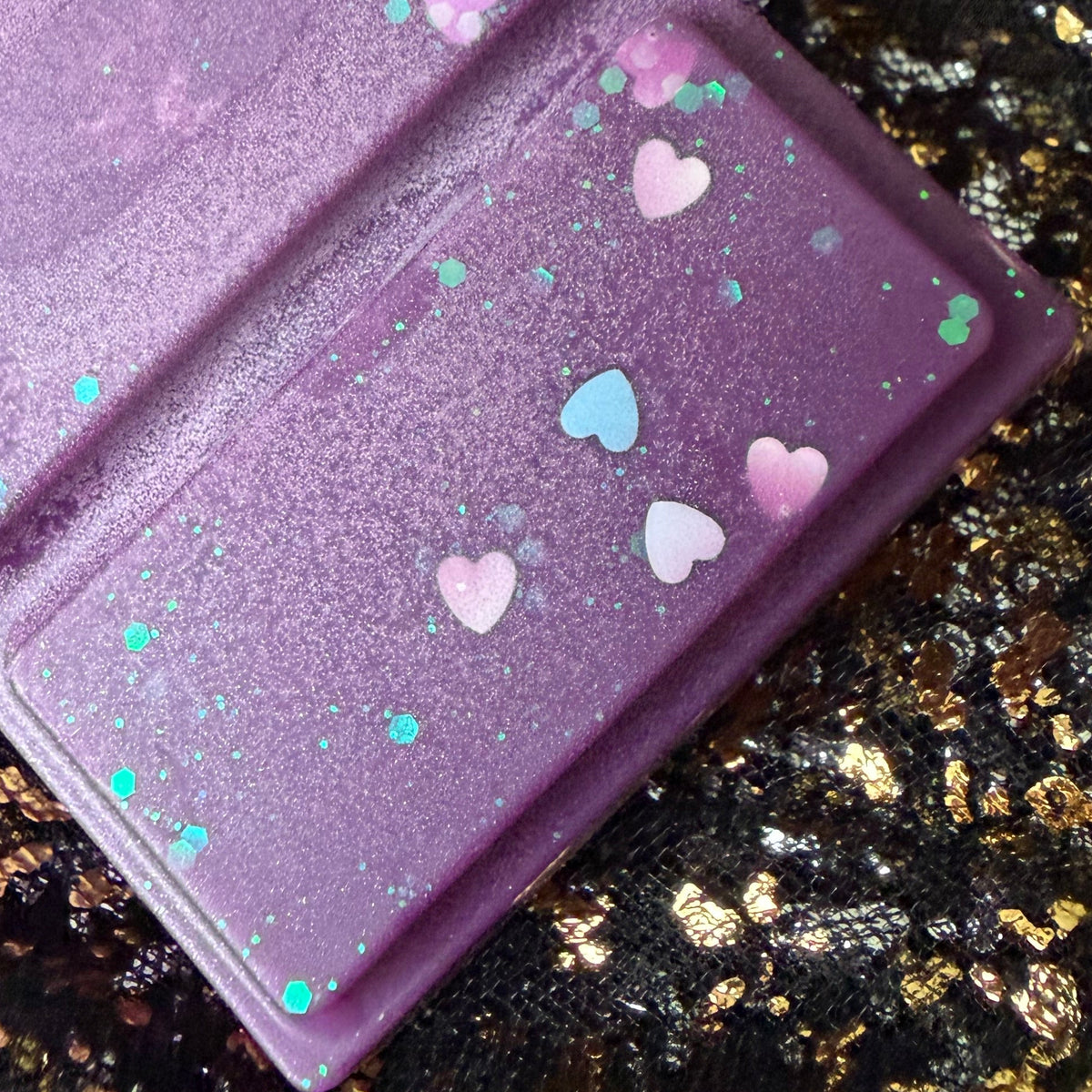 A close-up of a purple glittery soy wax melt decorated with small pink, white, and blue heart iridescent glitter, set against a black background with gold foil speckles.