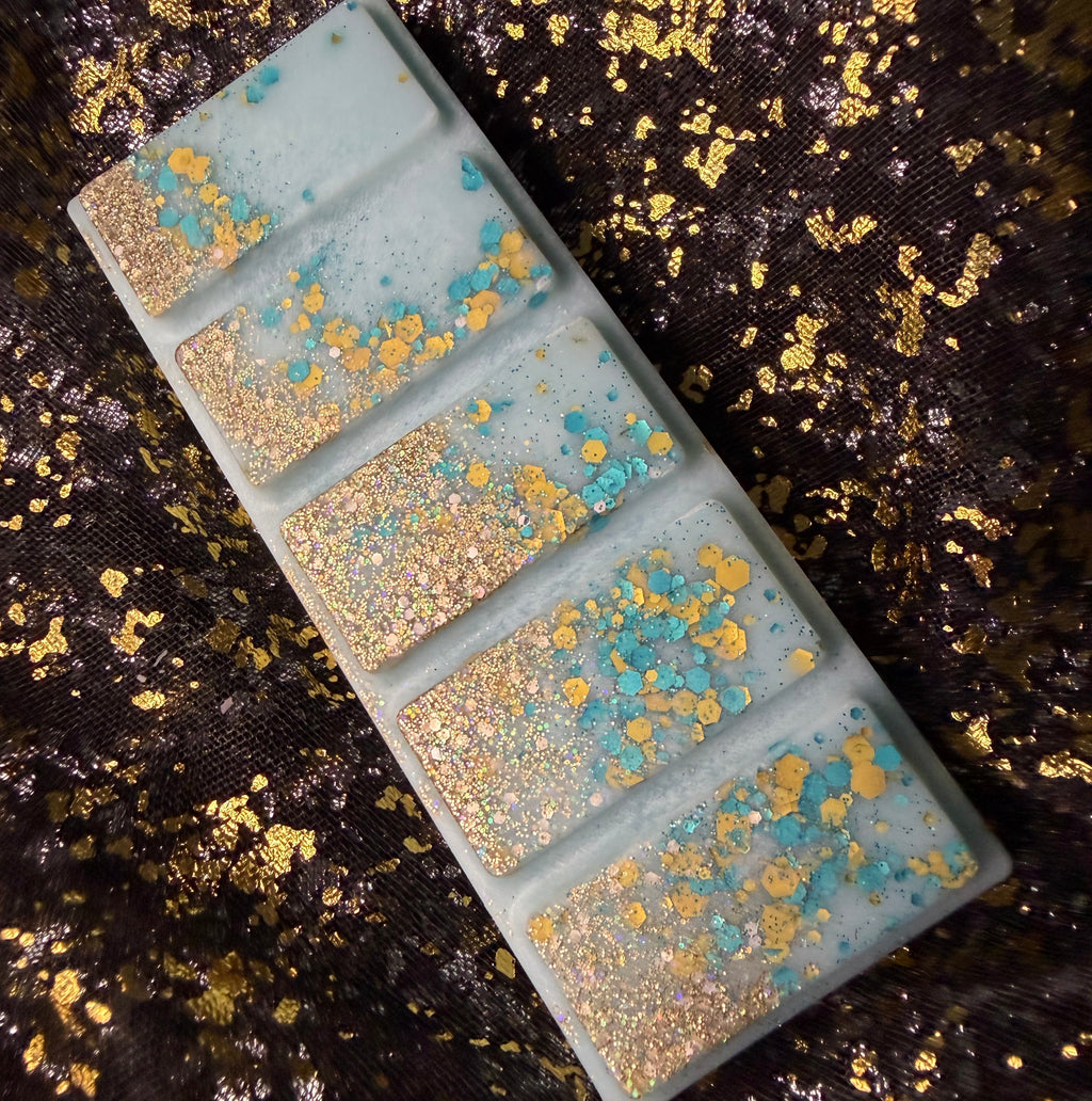 Secrets of the deep ocean scented soy wax melt bar showing five segments , featuring a pale blue-green base with shimmering gold foil and turquoise blue glitter, displayed diagonally on a dark textured fabric with gold flecks.