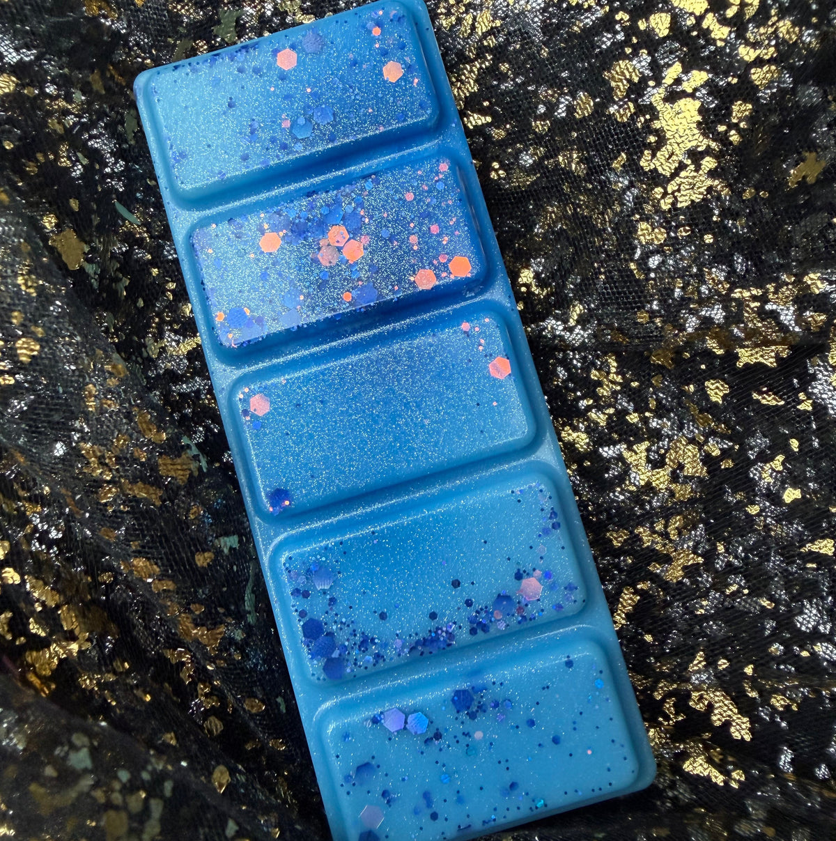 The Snowy Escape five-segment blue wax melt positioned sideways on black-and-gold fabric, showing its rectangular shape and purple glitter decoration.