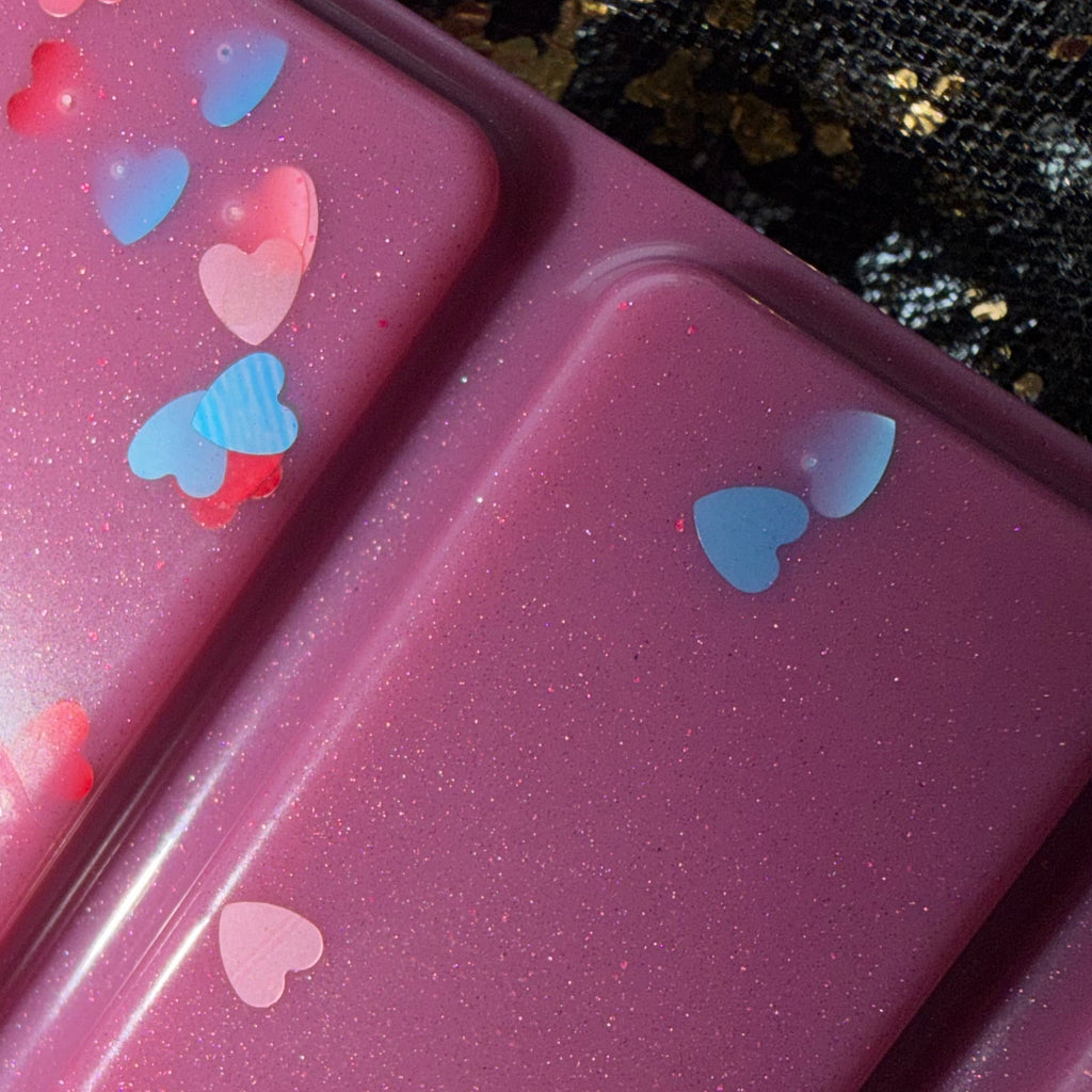 Close-up of two deep berry-purple Bryce After Dark wax melt segments decorated with red, pink and blue heart-shaped glitter against a black-and-gold fabric background.