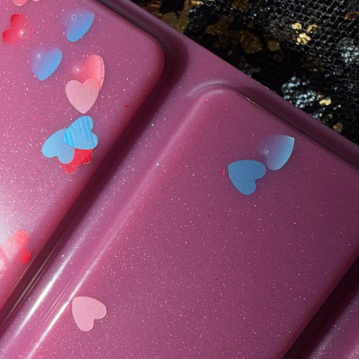 Close-up of two deep berry-purple Bryce After Dark wax melt segments decorated with red, pink and blue heart-shaped glitter against a black-and-gold fabric background.