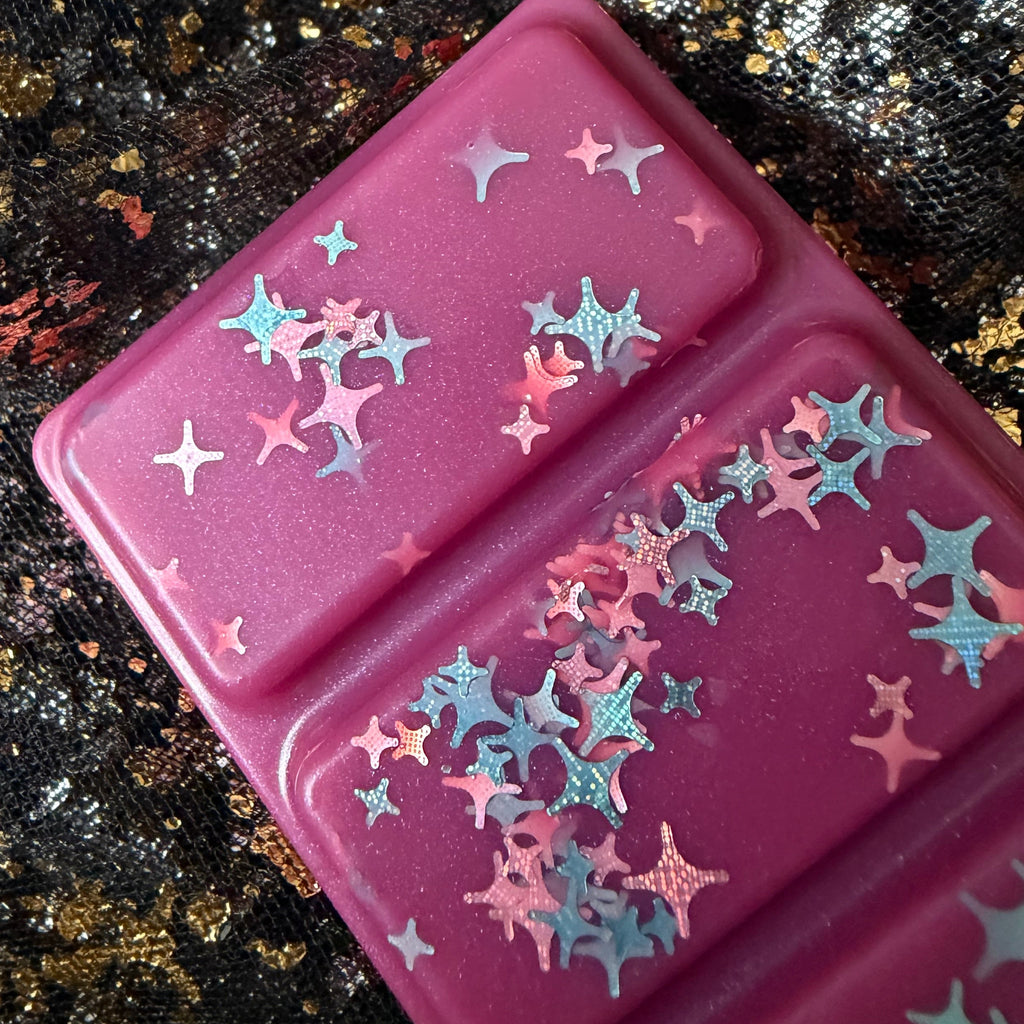 Close-up of two One More Chapter wax melt segments showing silver, blue, pink and iridescent star-shaped glitter against deep pink wax.