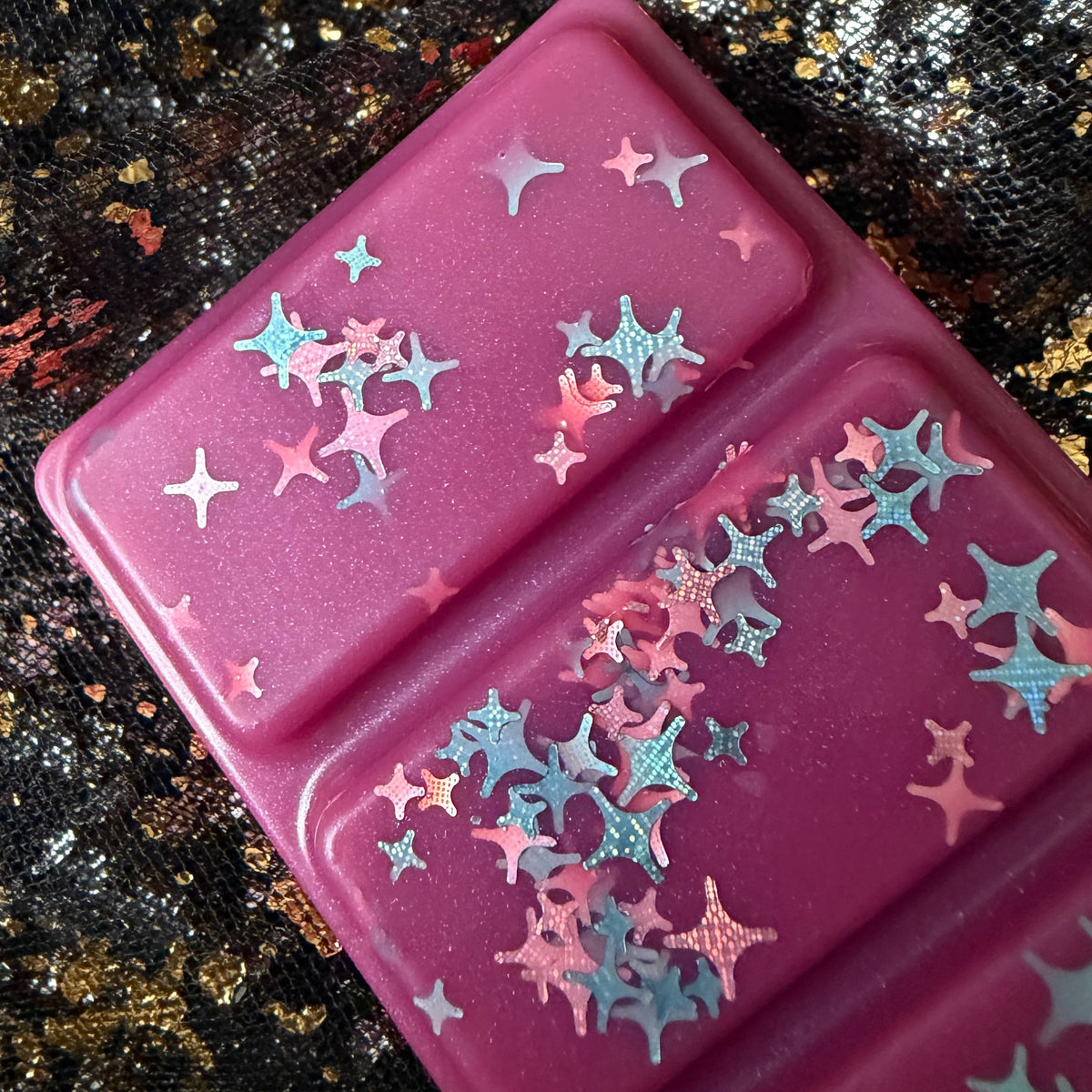 Close-up of two One More Chapter wax melt segments showing silver, blue, pink and iridescent star-shaped glitter against deep pink wax.