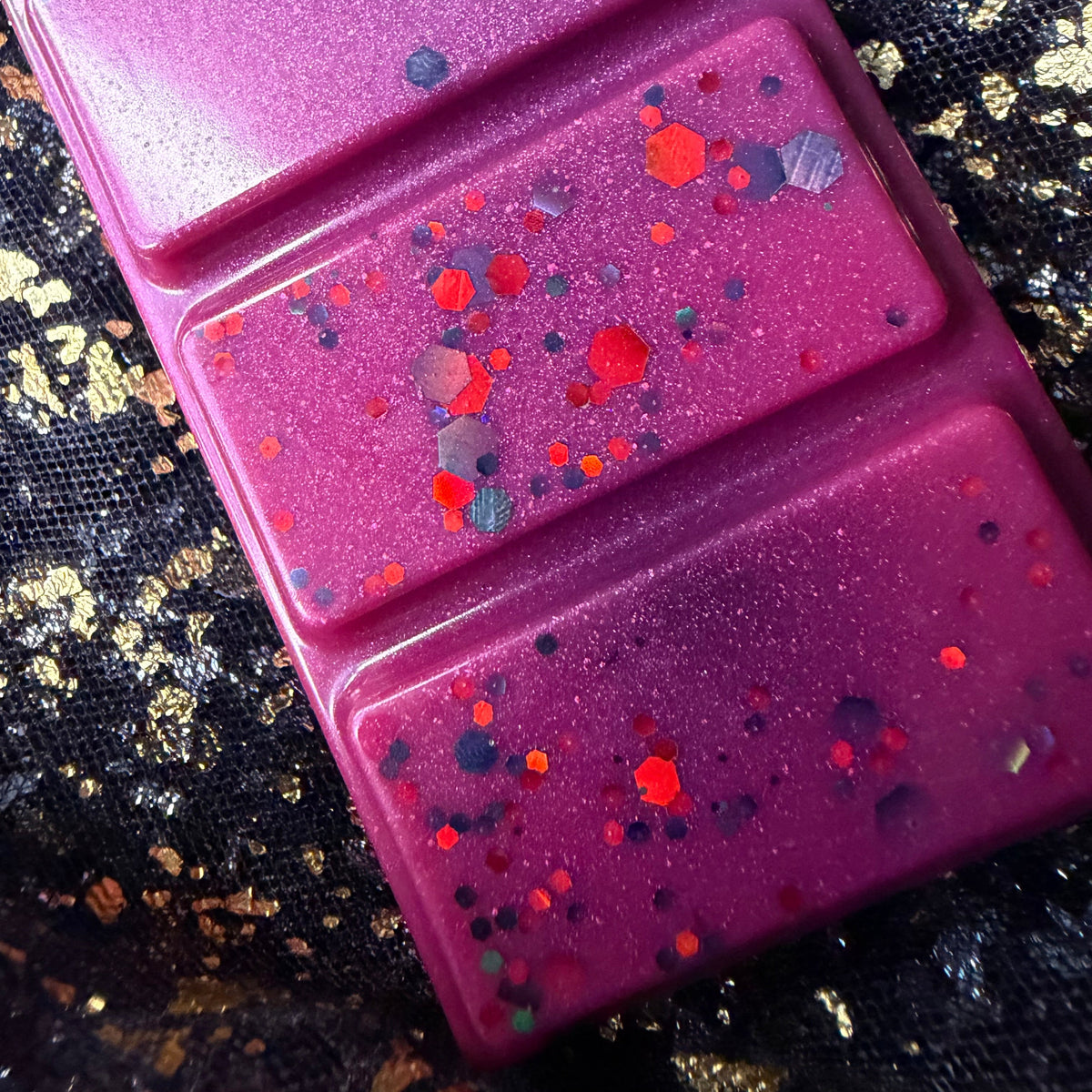 Close-up of two Shadowed Souls wax melt segments showing red, orange and charcoal glitter scattered across vivid magenta wax.