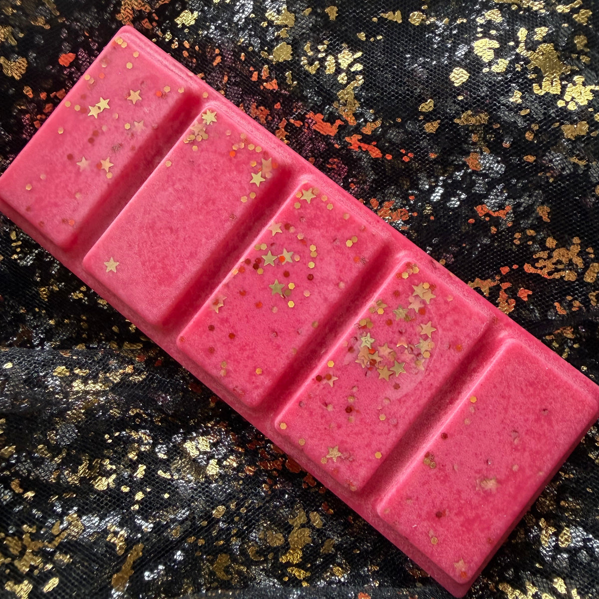 Full-length Love & Luck wax melt positioned diagonally on black-and-gold fabric, showing all five vivid red segments scattered with gold stars and fine glitter.