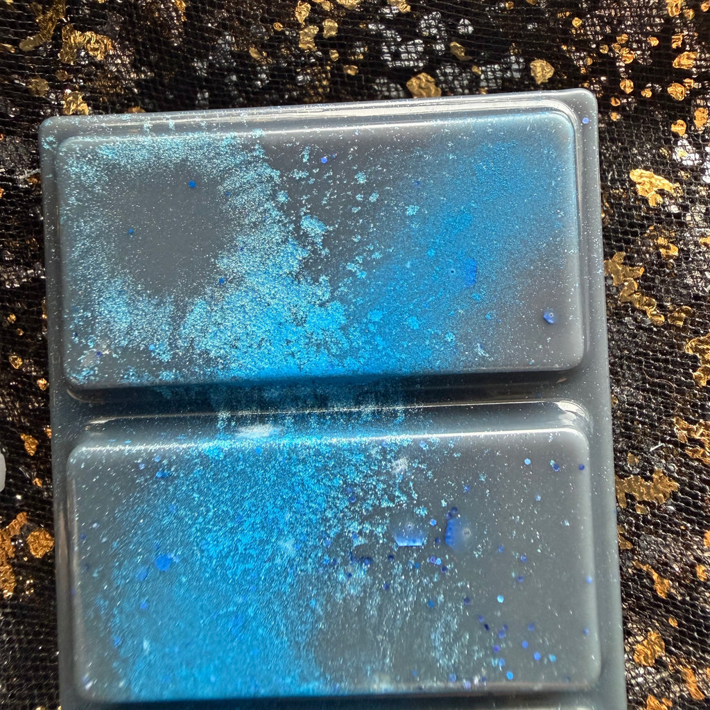 Close up of two Darcy Vega wax melt sections, showing bright blue colouring, pale turquoise shimmer and scattered blue hexagonal glitter against dark wax, bordered by black fabric with gold flecks.
