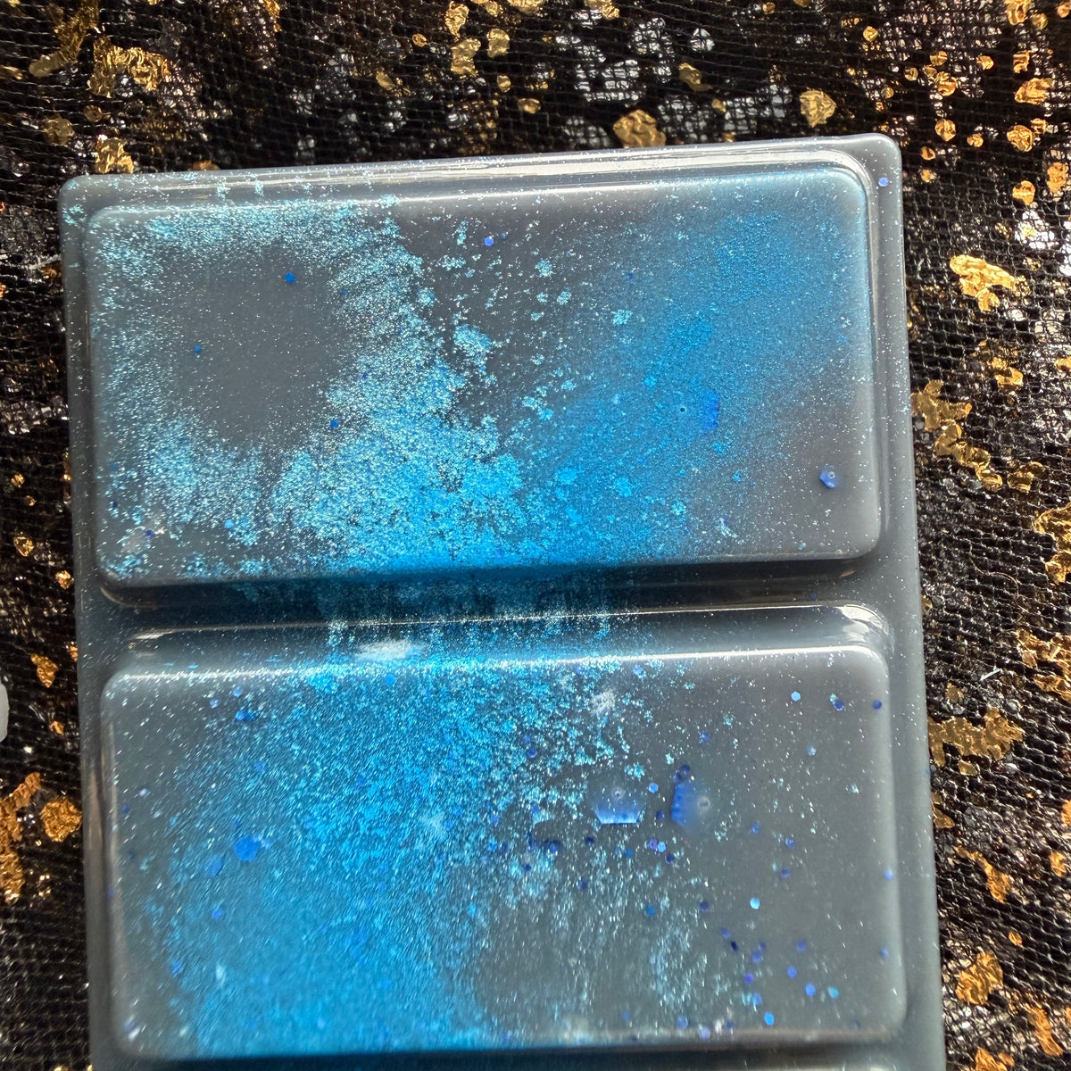 Close up of two Darcy Vega wax melt sections, showing bright blue colouring, pale turquoise shimmer and scattered blue hexagonal glitter against dark wax, bordered by black fabric with gold flecks.