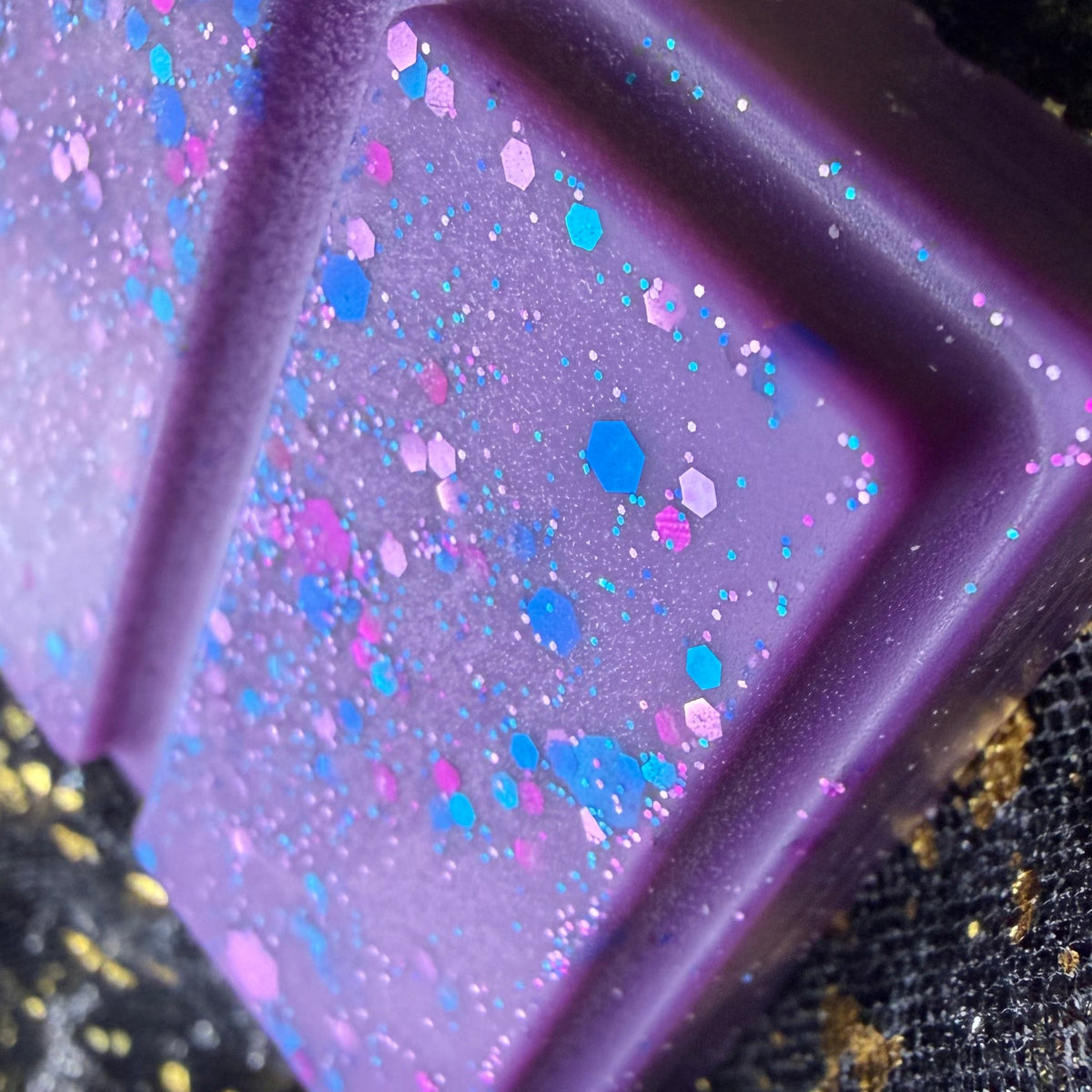 A close-up of the purple rectangular wax melt with embossed segments and holographic glitter speckles, resting on a black textured fabric with gold flecks.