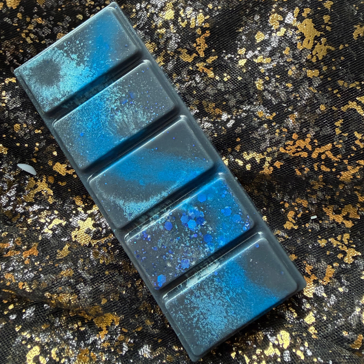 Darcy Vega five segment soy wax melt lying diagonally on black and gold fabric, showing a vivid blue streak, patches of turquoise shimmer and blue hexagonal glitter clustered on the fourth segment from the top.