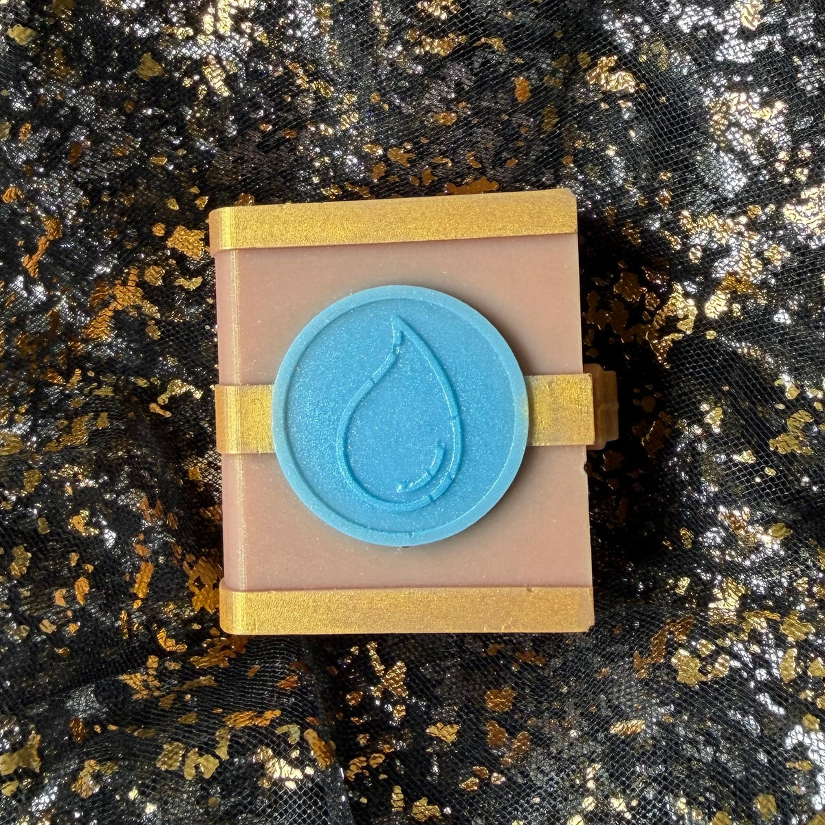 Full Water soy wax spellbook centred on black and gold fabric, with a dusty rose cover and shimmering blue circular water emblem.