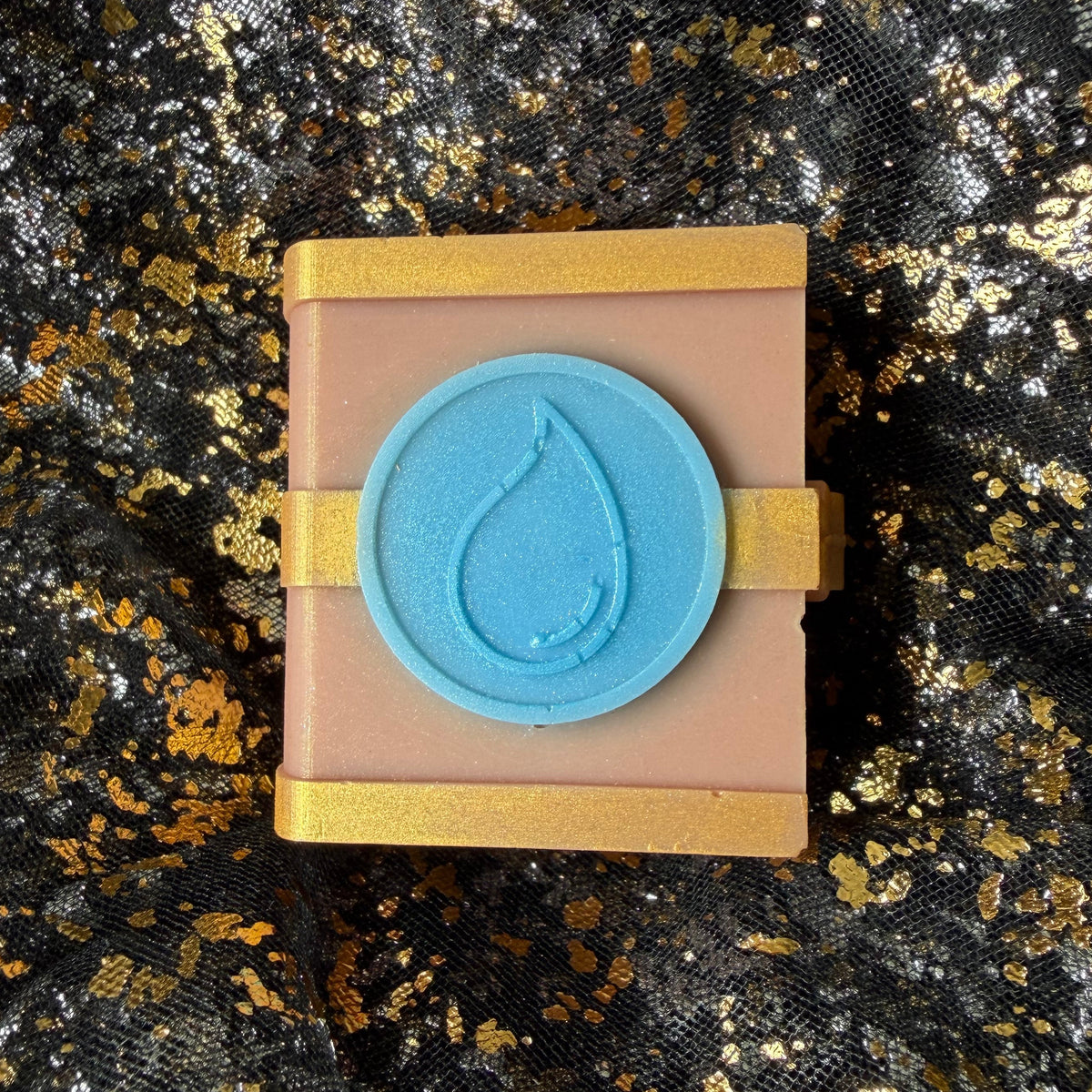 Water spellbook soy wax melt, citrus and aquatic long lasting scent viewed from above, with a shimmering blue droplet element and gold bands on its dusty rose cover.