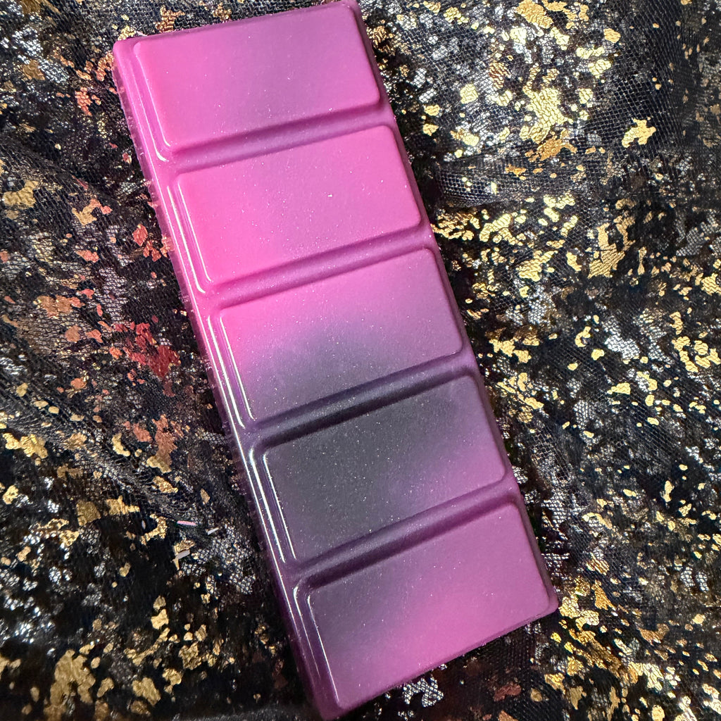 Full-length Joey wax melt positioned vertically on black-and-gold fabric, showing bright pink upper segments fading through charcoal grey into muted purple.