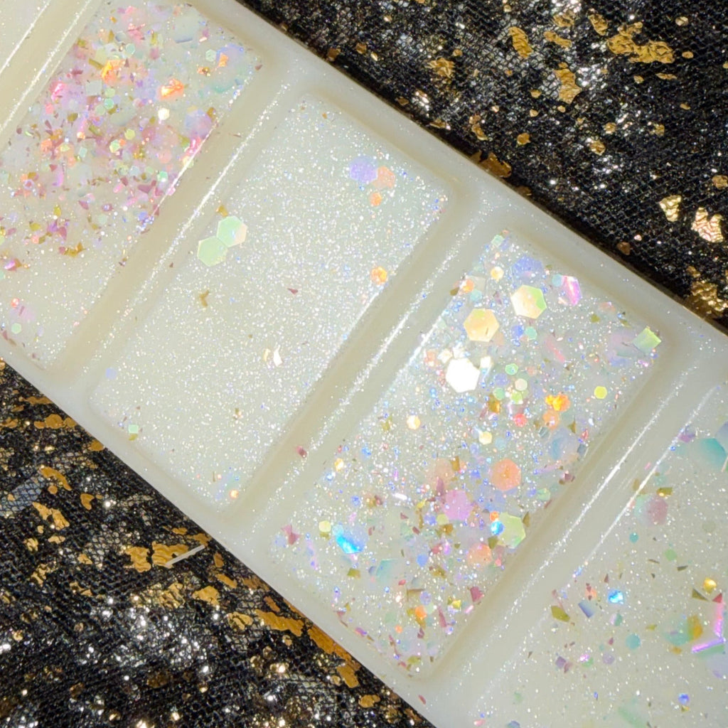 A rectangular translucent white soy wax melt scented baby powder with five sections, each filled with iridescent holographic glitter, displayed diagonally on a black sequined fabric background with gold flecks.