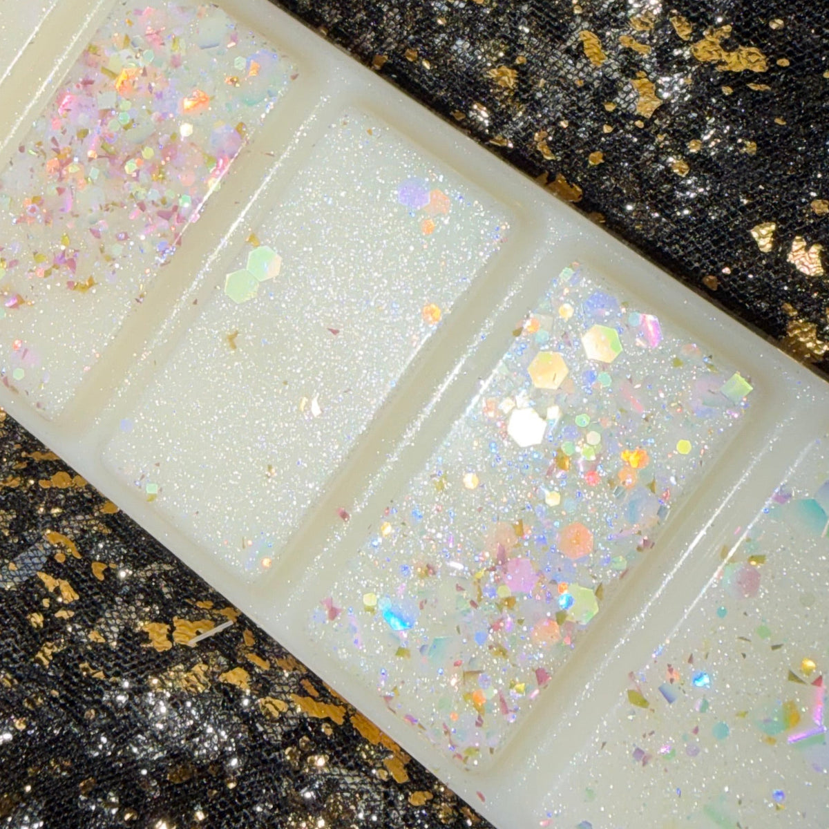 A rectangular translucent white soy wax melt scented baby powder with five sections, each filled with iridescent holographic glitter, displayed diagonally on a black sequined fabric background with gold flecks.