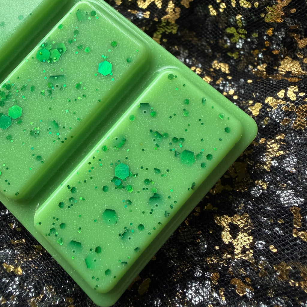 A close-up of Mirror Mirror a high-quality soy wax melt coloured green with handcrafted decorated green glitter and small translucent pieces, placed on a black sequined surface with gold flecks. 