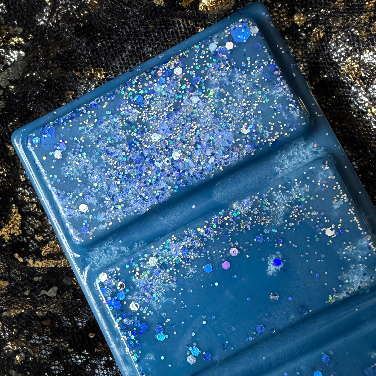 A rectangular navy blue ocean scented vegan soy wax melt, filled with shimmering blue and silver glitter, photographed diagonally on a black fabric background with gold flecks.