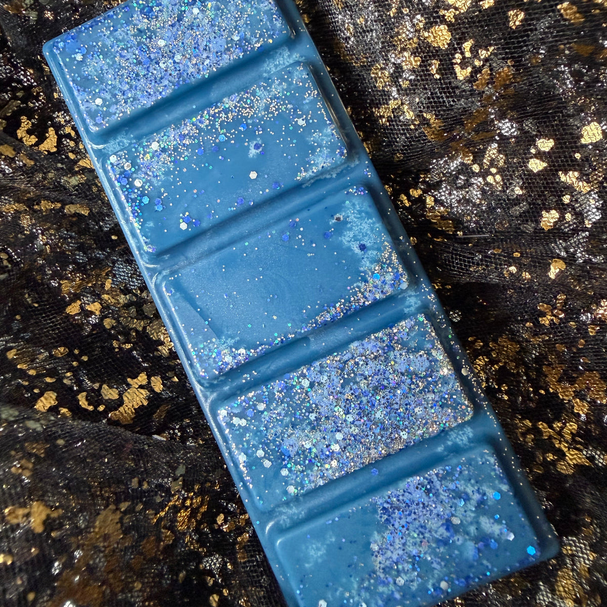 Deep blue Whitby Shadows wax bar displayed diagonally on black-and-gold fabric, with silver holographic glitter across all five segments.