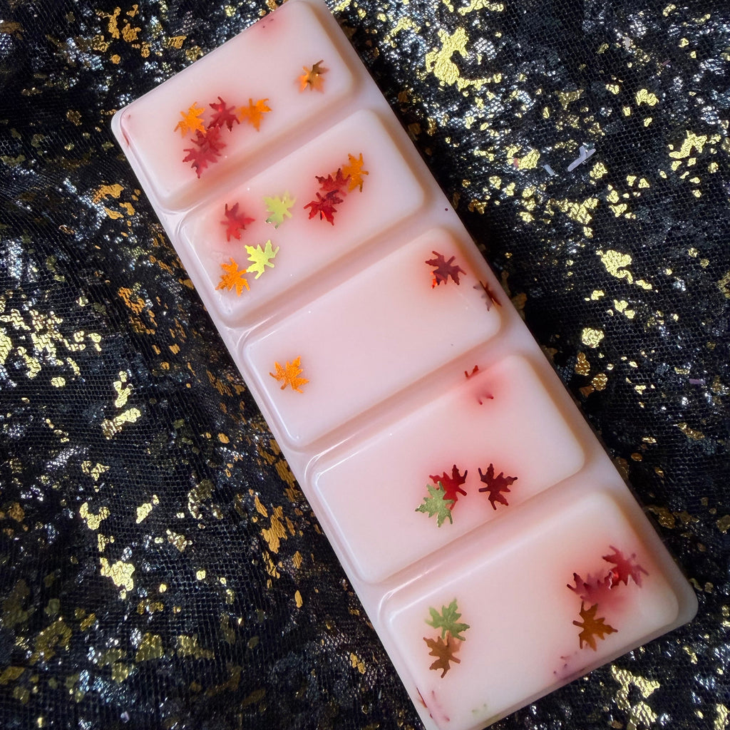 Willa’s Apple Pie scented wax melt, showing a sideways image of a rectangular five segment soy wax melt, each containing glitter shaped small autumn leaves in red, orange, and yellow, displayed diagonally on a black and gold glittery fabric background.