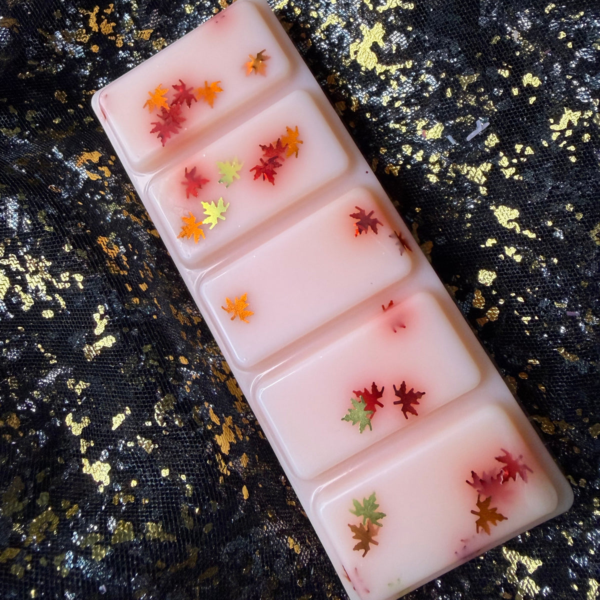 Willa’s Apple Pie scented wax melt, showing a sideways image of a rectangular five segment soy wax melt, each containing glitter shaped small autumn leaves in red, orange, and yellow, displayed diagonally on a black and gold glittery fabric background.