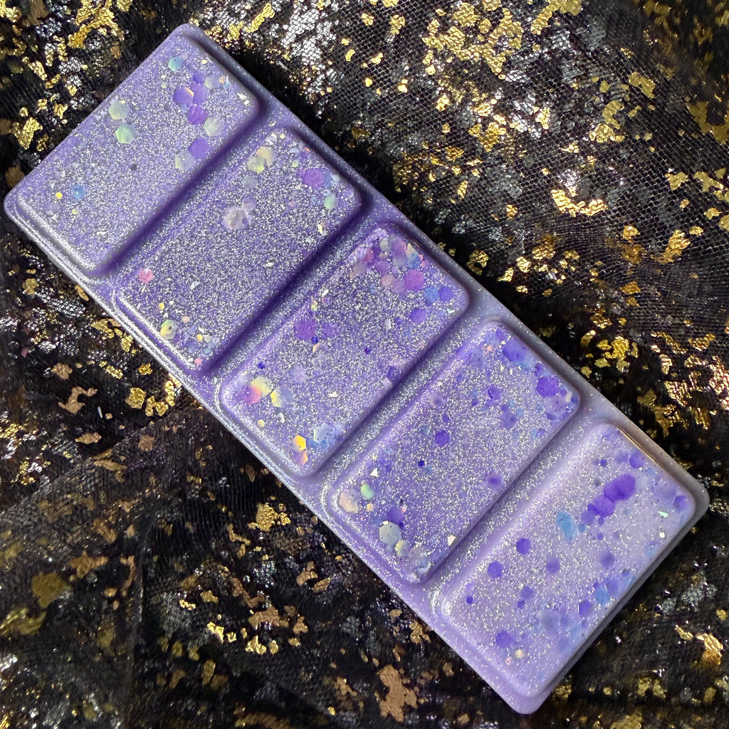 The Femme Fatale FMC-inspired high-quality soy wax melt removed from its clamshell and positioned at an angle, with citrus, jasmine, rose, amber and vanilla. Decorated with purple and iridescent glitter, resting on a black and gold textured fabric background. 