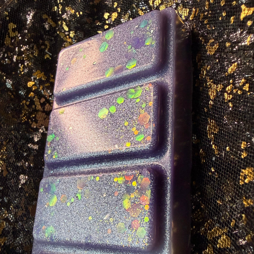 A close-up of The Femme Fatale - a fruity floral bookish wax melt.  rectangular wax melt with five grooved sections, featuring a dark purple base with vibrant lilac rainbow iridescent speckles of glitter, set against a black and gold textured background.