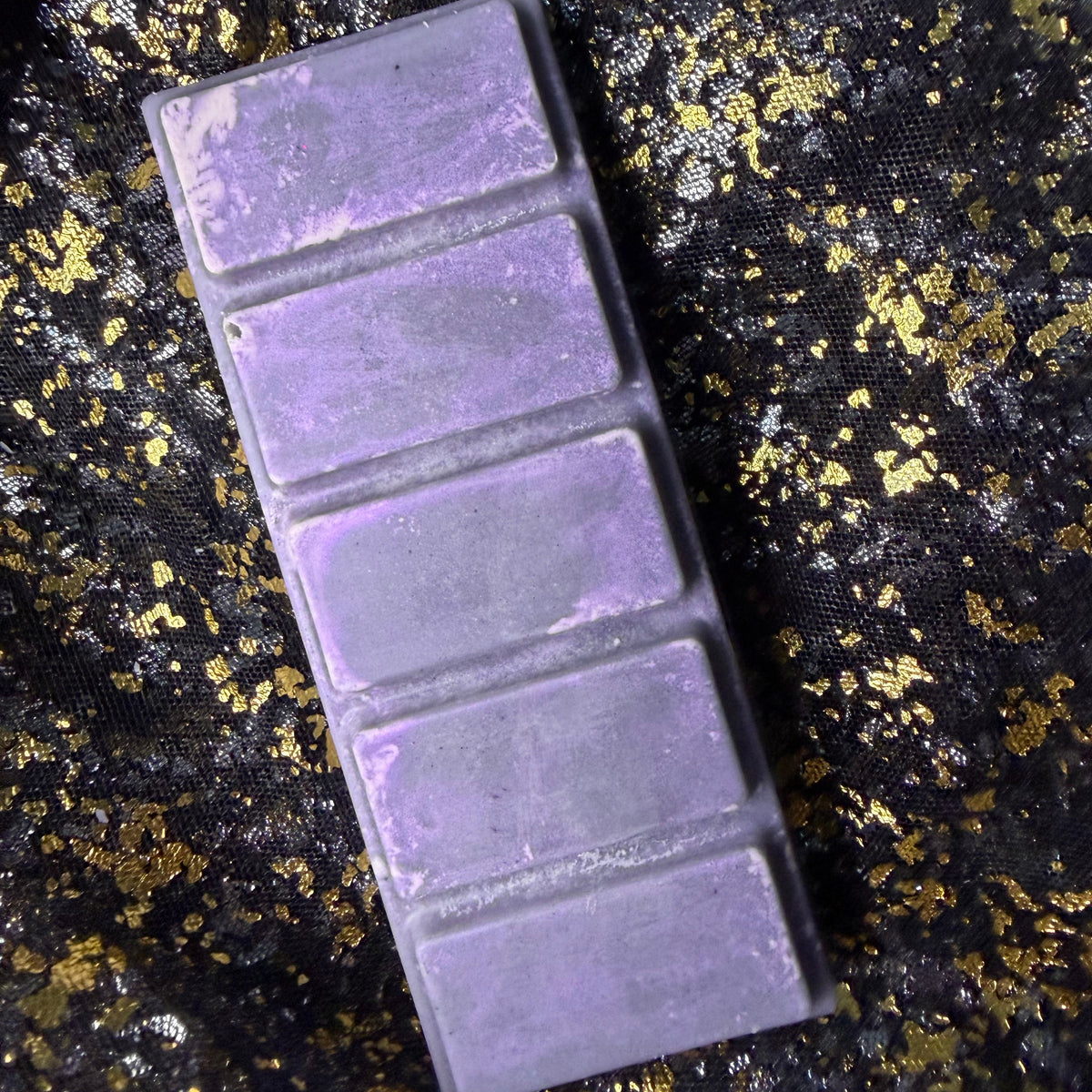 A rectangular purple wax melt bar with five segmented sections, smoky cedarwood scented soy wax melt shown closely outside its packaging. Placed diagonally on a textured black fabric background with scattered gold leaf.