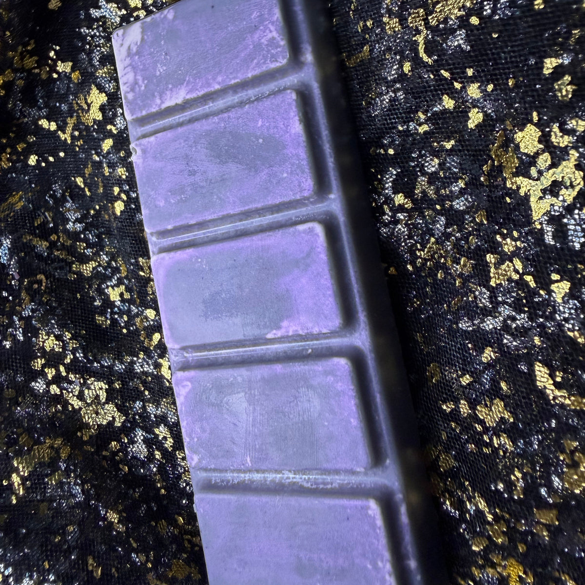 An angled view of the rectangular purple Smouldering Embers soy wax melt showing its segmented shape and finished surface with five segmented sections, placed diagonally on a black fabric background with gold glitter.
