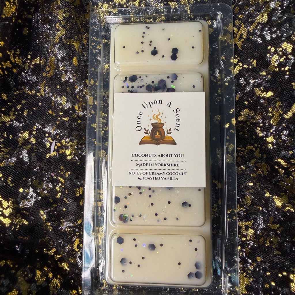 A clear plastic clamshell package containing four rectangular cream-colored wax melts with black speckles, featuring a white paper label that reads 'Once Upon A Sweet' and 'Coconutys About You'. The package is placed on a black sequined fabric background with gold glitter.