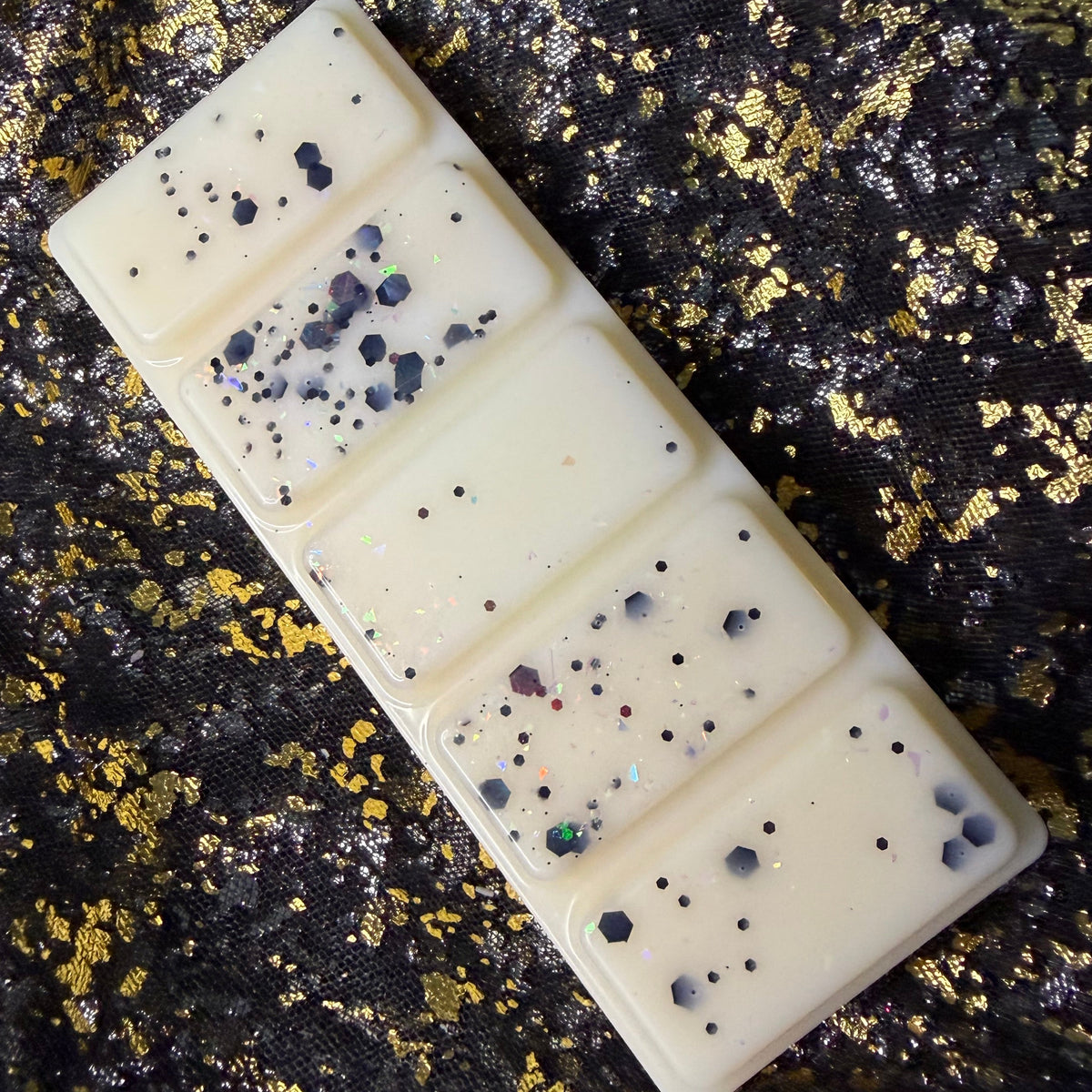 A rectangular cream-colored wax melt bar with black and multicolor glitter flecks, placed diagonally on a textured black and gold glittery fabric background.