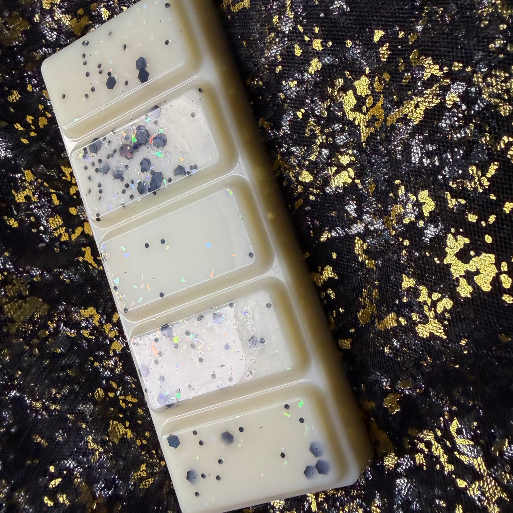 A rectangular cream-colored wax melt bar with five segmented sections, decorated with black speckles and iridescent glitter, displayed diagonally showing the glossy shine on a black fabric background with gold foil flakes.