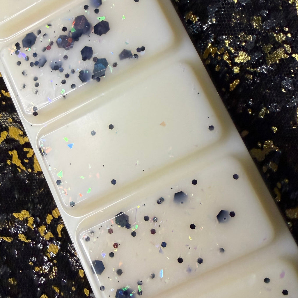 A close up of a rectangular cream-colored wax melt or with five rectangular sections, decorated with black glitter and iridescent flecks, shown on a black and gold textured background. 