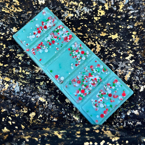 Christmas Tree wax melt displayed diagonally on black and gold fabric, showing a mint-green rectangular five-segment bar decorated with red, white and green festive confetti glitter.
