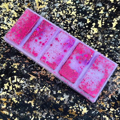 Cherries on Snow cherry and vanilla wax melt, showing an unwrapped pale lilac rectangular five-segment soy wax bar covered with fine and hexagonal bright pink glitter, displayed diagonally on black-and-gold fabric.
